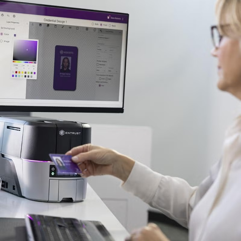 ID Card Software & Instant ID Support | ID CardSolutions - Person swiping a purple ID card into an Entrust card printer while a monitor on the desk shows design software.