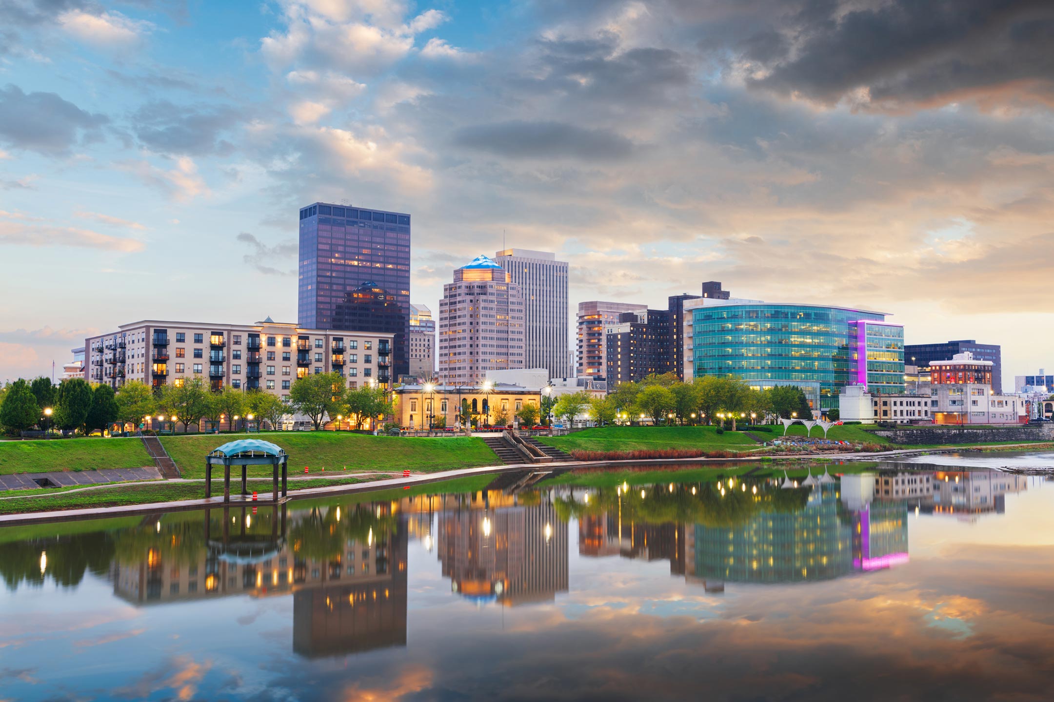 City skyline at sunset with modern glass buildings along a calm river and a grassy riverside park, reflecting in the water.